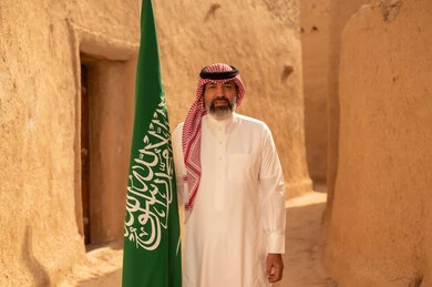 Celebrating Saudi National Day, September 23, gestures of pride and joy in belonging to the homeland, a smiling Saudi Arabian Gulf man wearing a traditional shemagh and thobe standing in a mud house holding the Kingdom's flag, the day we began in 1727 AD, the art of ancient architecture and building mud houses, Flag Day, March 11