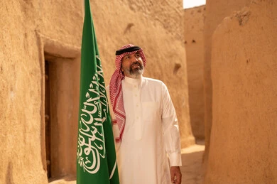 إيماءات الفخر و السرور بالانتماء للوطن, رجل عربي خليجي سعودي مبتسم يرتدي الشماغ و الثوب التقليدي يقف في بيت طيني ويحمل بيده علم المملكة، يوم بدينا 1727 م، فن العمارة القديم وبناء البيوت الطينية، يوم العلم 11 مارس، إحياء ذكرى اليوم الوطني السعودي 23 سبتمبر