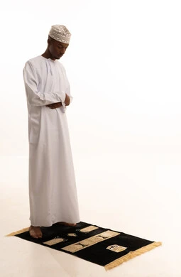 بورتريه لشاب عربي عماني خليجي اسمر يرتدي الدشداشة والكمة يقف على السجادة و يؤدي فريضة الصلاة ، آداء صلاة التراويح ، الدعاء والتضرع الى الله، أذكار وأدعية وفرائض، الركوع والسجود والخشوع لله، خلفية بيضاء.
