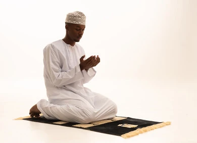 Bowing, prostration, and humility before God, a portrait of a young Arab Saudi Gulf man with a dark complexion wearing traditional attire, standing on a prayer rug and performing the prayer obligation, performing Taraweeh prayer, supplication and pleading to God, remembrances, prayers, and obligations, with a white background.