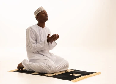 Bowing, prostration, and humility before God, a portrait of a young Arab Saudi Gulf man with a dark complexion wearing traditional attire, standing on a prayer rug and performing the prayer obligation, performing Taraweeh prayer, supplication and pleading to God, remembrances, prayers, and obligations, with a white background.