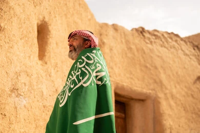 إحياء ذكرى اليوم الوطني السعودي 23 سبتمبر, إيماءات الفخر و السرور بالانتماء للوطن, رجل عربي خليجي سعودي مبتسم يرتدي الشماغ و الثوب التقليدي يقف في بيت طيني ويحمل بيده علم المملكة، يوم بدينا 1727 م، فن العمارة القديم وبناء البيوت الطينية، يوم العلم 11 مارس
