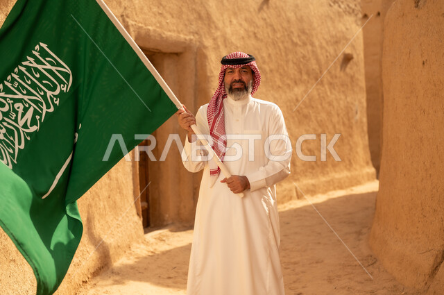 إحياء ذكرى اليوم الوطني السعودي 23 سبتمبر, إيماءات الفخر و السرور بالانتماء للوطن, رجل عربي خليجي سعودي مبتسم يرتدي الشماغ و الثوب التقليدي يقف في بيت طيني ويحمل بيده علم المملكة، يوم بدينا 1727 م، فن العمارة القديم وبناء البيوت الطينية، يوم العلم 11 مارس