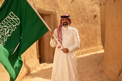 Celebrating Saudi National Day, September 23, gestures of pride and joy in belonging to the homeland, a smiling Saudi Arabian Gulf man wearing a traditional shemagh and thobe standing in a mud house holding the Kingdom's flag, the day we began in 1727 AD, the art of ancient architecture and building mud houses, Flag Day, March 11