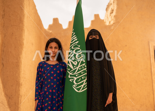 يوم العلم 11 مارس، ذكرى تأسيس الدولة السعودية الاولى 22 فبراير، إمرأة عربيه خليجية سعودية مبتسمة ترتدي البرقع والعباءة تمسك بيدها العلم تقف في منزل طيني برفقة ابنتها، اليوم الوطني السعودي 23 سبتمبر، المحافظة على التراث السعودي، تربيه الأبناء على حب الوطن