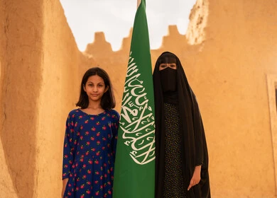 يوم العلم 11 مارس، ذكرى تأسيس الدولة السعودية الاولى 22 فبراير، إمرأة عربيه خليجية سعودية مبتسمة ترتدي البرقع والعباءة تمسك بيدها العلم تقف في منزل طيني برفقة ابنتها، اليوم الوطني السعودي 23 سبتمبر، المحافظة على التراث السعودي، تربيه الأبناء على حب الوطن