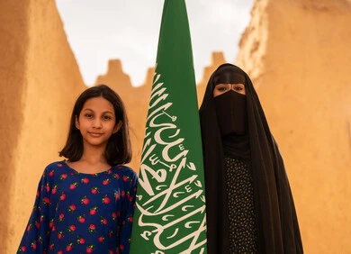 تربيه الأبناء على حب الوطن، يوم العلم 11 مارس، ذكرى تأسيس الدولة السعودية الاولى 22 فبراير، إمرأة عربيه خليجية سعودية مبتسمة ترتدي البرقع والعباءة تمسك بيدها العلم تقف في منزل طيني برفقة ابنتها، اليوم الوطني السعودي 23 سبتمبر، المحافظة على التراث السعودي