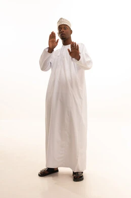 Protecting oneself from external harm, a request to stop and refrain from doing something, terror and panic and dread of something, a portrait of a young Arab Gulf Omani man with a dark complexion wearing a dishdasha and kumma raising his hand upwards in gestures of fear, white background.