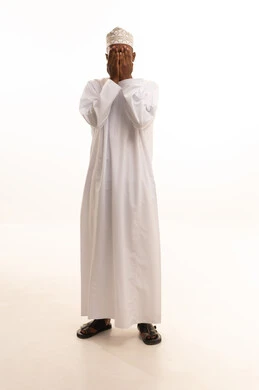 A portrait of a young Arab Gulf Omani man with a dark complexion wearing a dishdasha and a kumma, hiding his face with his hands. The gestures of his hands indicate hiding; a shy man concealing his face with his hands, preventing sight of something, the concept of not seeing, with a white background.