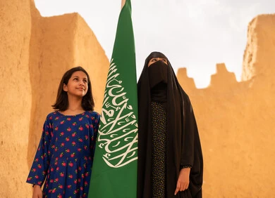 تربيه الأبناء على حب الوطن، يوم العلم 11 مارس، ذكرى تأسيس الدولة السعودية الاولى 22 فبراير، إمرأة عربيه خليجية سعودية مبتسمة ترتدي البرقع والعباءة تمسك بيدها العلم تقف في منزل طيني برفقة ابنتها، اليوم الوطني السعودي 23 سبتمبر، المحافظة على التراث السعودي