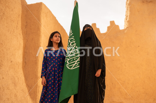 يوم العلم 11 مارس، ذكرى تأسيس الدولة السعودية الاولى 22 فبراير، إمرأة عربيه خليجية سعودية مبتسمة ترتدي البرقع والعباءة تمسك بيدها العلم تقف في منزل طيني برفقة ابنتها، اليوم الوطني السعودي 23 سبتمبر، المحافظة على التراث السعودي، تربيه الأبناء على حب الوطن