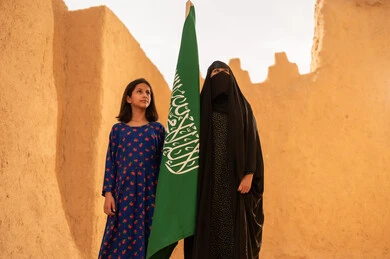 يوم العلم 11 مارس، ذكرى تأسيس الدولة السعودية الاولى 22 فبراير، إمرأة عربيه خليجية سعودية مبتسمة ترتدي البرقع والعباءة تمسك بيدها العلم تقف في منزل طيني برفقة ابنتها، اليوم الوطني السعودي 23 سبتمبر، المحافظة على التراث السعودي، تربيه الأبناء على حب الوطن