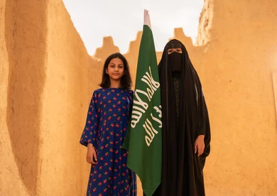 تربيه الأبناء على حب الوطن، يوم العلم 11 مارس، ذكرى تأسيس الدولة السعودية الاولى 22 فبراير، إمرأة عربيه خليجية سعودية مبتسمة ترتدي البرقع والعباءة تمسك بيدها العلم تقف في منزل طيني برفقة ابنتها، اليوم الوطني السعودي 23 سبتمبر، المحافظة على التراث السعودي