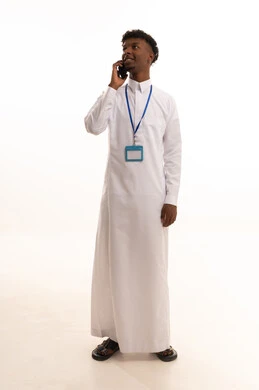 Making a phone call, using modern technologies, a portrait of a young Arab Gulf Saudi man with a dark complexion wearing a white thobe using a mobile phone, wearing a personal badge, a personal identification card with the employee's name, a blank white rectangular mock-up, white background.
