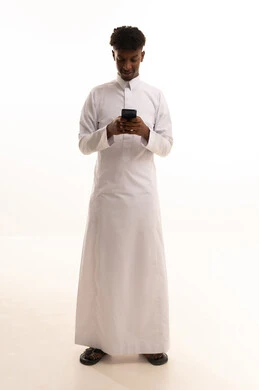 The use of modern technologies, browsing social media sites via mobile, completing work tasks remotely, a portrait of a young Arab Gulf Saudi man with a dark complexion wearing a white thobe using a mobile phone, white background.