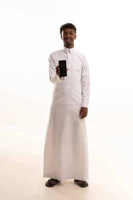 The use of modern technologies, browsing social media sites via mobile, completing work tasks remotely, a portrait of a young Arab Gulf Saudi man with a dark complexion wearing a white thobe using a mobile phone, white background.