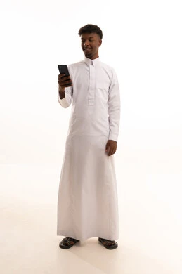 A portrait of a young Arab Gulf Saudi man with a tan wearing a white thobe, using a mobile phone, utilizing modern technologies, browsing social media on his phone, completing work tasks remotely, with a white background.
