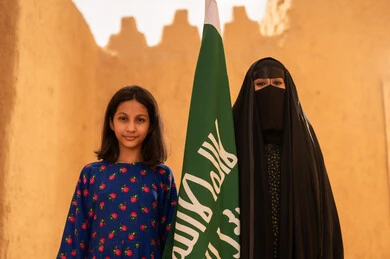 Raising children to love their country, Flag Day March 11, the anniversary of the founding of the first Saudi state February 22, a smiling Saudi Gulf Arab woman wearing a burqa and abaya holding a flag in her hand, standing in a mud house with her daughter, Saudi National Day September 23, preserving Saudi heritage