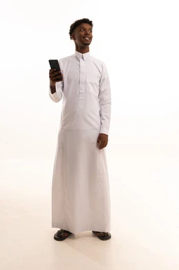 The use of modern technologies, browsing social media sites via mobile, completing work tasks remotely, a portrait of a young Arab Gulf Saudi man with a dark complexion wearing a white thobe using a mobile phone, white background.