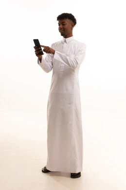 تصفح مواقع التواصل الاجتماعي عبر الجوال، استخدام التقنيات الحديثة، إنجاز مهام العمل عن بعد، بورتريه لشاب عربي خليجي سعودي اسمر يرتدي الثوب الابيض يقوم باستخدام الهاتف المحمول، خلفية بيضاء