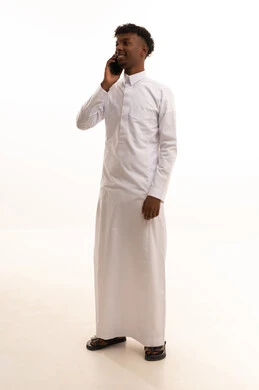 The use of modern technologies, browsing social media sites via mobile, completing work tasks remotely, a portrait of a young Arab Gulf Saudi man with a dark complexion wearing a white thobe using a mobile phone, white background.