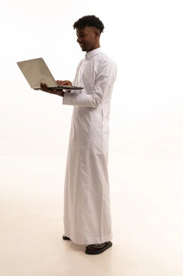 Remote work, using a laptop, a portrait of a young Arab Gulf Saudi man with a dark complexion wearing a white thobe browsing the internet, using modern technology devices, conducting a live broadcast via the laptop, white background.