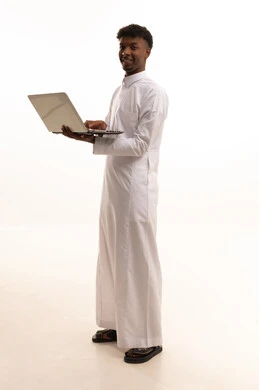 إستخدام جهاز الحاسوب المحمول،بورتريه لشاب عربي خليجي سعودي اسمر يرتدي الثوب الابيض يتصفح الانترنت ، إستخدام اجهزة تقنية حديثة ، العمل عن بعد ، اجراء بث مباشر عبر اللابتوب، خلفية بيضاء