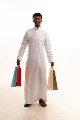 الاستفادة من العروضات والتخفيضات، المتعة في التسوق واقتناء المستلزمات والهدايا، بورتريه لشاب عربي خليجي سعودي اسمر يرتدي الثوب الابيض  يحمل بيده أكياس ورقية ملونة بإيماءات السرور والسعادة، الجسم بالطول الكامل، خلفية بيضاء