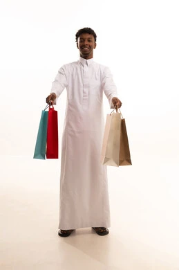 المتعة في التسوق واقتناء المستلزمات والهدايا، الاستفادة من العروضات والتخفيضات، بورتريه لشاب عربي خليجي سعودي اسمر يرتدي الثوب الابيض  يحمل بيده أكياس ورقية ملونة بإيماءات السرور والسعادة، الجسم بالطول الكامل، خلفية بيضاء