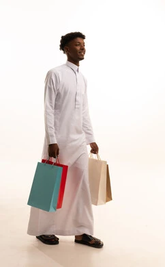A portrait of a young Arab Gulf Saudi man with a tan wearing a white thobe, holding colorful paper bags in his hand with gestures of joy and happiness, enjoying shopping and acquiring supplies and gifts, taking advantage of offers and discounts, full-body shot, white background.