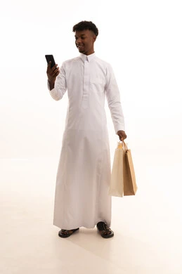 Making a phone call via mobile, buying supplies and gifts, communicating with family and friends, a portrait of a young Arab Saudi Gulf man with a dark complexion wearing traditional attire holding shopping bags and a mobile phone in his hands, a full-body image, white background.