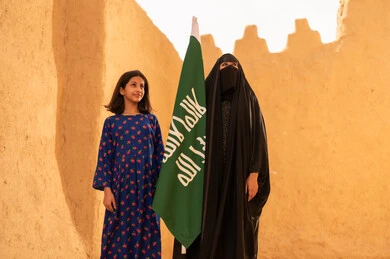 Raising children to love their country, Flag Day March 11, the anniversary of the founding of the first Saudi state February 22, a smiling Saudi Gulf Arab woman wearing a burqa and abaya holding a flag in her hand, standing in a mud house with her daughter, Saudi National Day September 23, preserving Saudi heritage