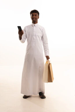 التواصل مع العائلة و الأصدقاء , إجراء مكالمة هاتفية عبر الجوال , شراء المستلزمات و الهدايا , بورتريه لشاب عربي سعودي خليجي اسمر يرتدي الثوب التقليدي يحمل بيديه أكياس التسوق والهاتف المحمول, صورة للجسم بالطول الكامل , خلفية بيضاء
