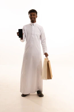 Buying supplies and gifts, making a phone call via mobile, communicating with family and friends, a portrait of a young Arab Saudi Gulf man with a dark complexion wearing traditional attire holding shopping bags and a mobile phone in his hands, a full-body image, white background.
