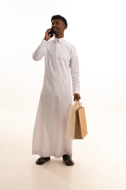 إجراء مكالمة هاتفية عبر الجوال , شراء المستلزمات و الهدايا , التواصل مع العائلة و الأصدقاء , بورتريه لشاب عربي سعودي خليجي اسمر يرتدي الثوب التقليدي يحمل بيديه أكياس التسوق والهاتف المحمول, صورة للجسم بالطول الكامل , خلفية بيضاء
