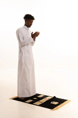 بورتريه لشاب عربي سعودي خليجي اسمر يرتدي الثوب التقليدي يقف على السجادة و يؤدي فريضة الصلاة ، آداء صلاة التراويح ، الدعاء والتضرع الى الله، أذكار وأدعية وفرائض، الركوع والسجود والخشوع لله، خلفية بيضاء.