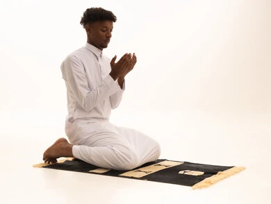 Bowing, prostration, and humility before God, a portrait of a young Arab Saudi Gulf man with a dark complexion wearing traditional attire, standing on a prayer rug performing the prayer obligation, performing Taraweeh prayer, supplication and pleading to God, remembrances, prayers, and obligations, with a white background.