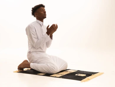 Bowing, prostration, and humility before God, a portrait of a young Arab Saudi Gulf man with a dark complexion wearing traditional attire, standing on a prayer rug performing the prayer obligation, performing Taraweeh prayer, supplication and pleading to God, remembrances, prayers, and obligations, with a white background.