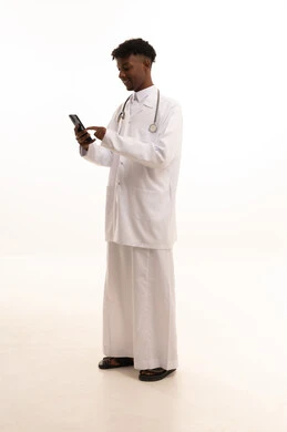 The use of technology and modern techniques, making a phone call, working in the Saudi health sector, providing services to patients with efficiency, the concept of medicine and healthcare, a portrait of a smiling Saudi Gulf doctor with a dark complexion wearing traditional attire and a white coat holding a mobile phone in his hand with gestures of joy, white background.