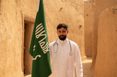 Saudi medical professions and jobs, the concept of working in the medical sector, a Saudi Gulf Arab doctor wearing the traditional medical coat and stethoscope standing in a mud house and holding the flag of the Kingdom, commemorating Saudi National Day, September 23, the day we started in 1727 AD, celebrating Flag Day, March 11