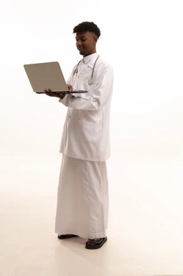مفهوم الطب والرعاية الصحية ، بورتريه لطبيبب سعودي خليجي اسمر مبتسم يرتدي الزي التقليدي والمعطف الابيض يمسك الحاسوب المحمول بيده بايماءات السرور، استخدام التكنولوجيا والتقنيات الحديثة، اجراء مكالمات الفيديو بتعابير الترحيب، العمل في القطاع الصحي السعودي، تقديم الخدمات للمرضى بكل كفاءه، ، خلفية بيضاء