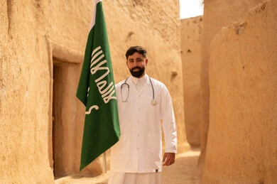 Celebrating Flag Day on March 11, Saudi medical professions and jobs, the concept of working in the medical sector, a Saudi Gulf Arab doctor wearing the traditional medical coat and stethoscope standing in a mud house holding the Kingdom's flag, commemorating Saudi National Day on September 23, the day we began in 1727 AD