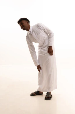 بورتريه لشاب عربي خليجي سعودي اسمر يرتدي الثوب الأبيض يضع يده على رجله بايماءات الألم والتعب والأرهاق، طلب المساعده والحاجه للعلاج، التهاب العضلات والمفاصل، الأوجاع والآلام العصبية،خلفية بيضاء.