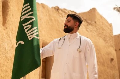 مهن ووظائف سعودية طبية، مفهوم العمل في القطاع الطبي، طبيب عربي خليجي سعودي يرتدي الثوب التقليدي المعطف الطبي والسماعات يقف في بيت طيني ويحمل بيده علم المملكة، إحياء ذكرى اليوم الوطني السعودي 23 سبتمبر، يوم بدينا 1727 م، الإحتفال بيوم العلم 11 مارس