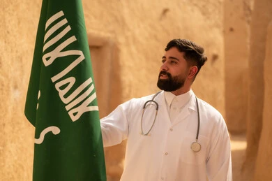 الإحتفال بيوم العلم 11 مارس، مهن ووظائف سعودية طبية، مفهوم العمل في القطاع الطبي، طبيب عربي خليجي سعودي يرتدي الثوب التقليدي المعطف الطبي والسماعات يقف في بيت طيني ويحمل بيده علم المملكة، إحياء ذكرى اليوم الوطني السعودي 23 سبتمبر، يوم بدينا 1727 م
