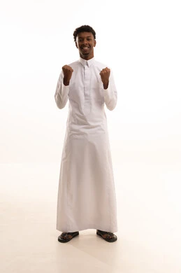 Gestures of victory and triumph, a portrait of a young Arab Gulf Saudi man with a dark complexion wearing a white thobe, raising his hands with expressions of interaction and enthusiasm, engaged in cheering for a football team, white background.