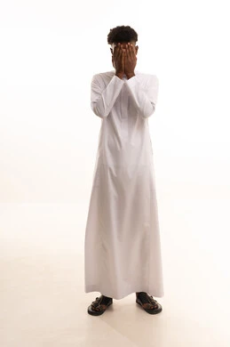 A portrait of a young Arab Gulf Saudi man with a dark complexion wearing a white thobe, hiding his face with his hands. The gestures of his hands indicate hiding; a shy man concealing his face with his hands, preventing himself from looking at something, the concept of not seeing, with a white background.