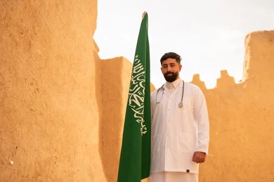 Saudi medical professions and jobs, the concept of working in the medical sector, a Saudi Gulf Arab doctor wearing the traditional medical coat and stethoscope standing in a mud house and holding the flag of the Kingdom, commemorating Saudi National Day, September 23, the day we started in 1727 AD, celebrating Flag Day, March 11