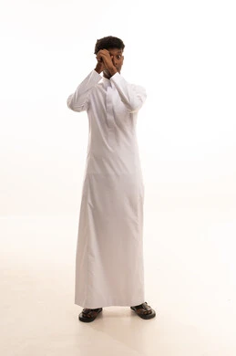Signs of focusing on something, searching and inquiring about a specific matter, feeling happy to discover the truths, a portrait of a young Arab Gulf Saudi man with a dark complexion wearing a white thobe, placing his hand over his eye to scrutinize and look at something specific, with a white background.
