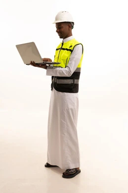 بورتريه لمهندس عربي خليجي سعودي اسمر يرتدي الثوب التقليدي والسترة الخاصة وخوذة الحماية يقف وبيده الحاسوب المحمول ، استخدام التكنولوجيا والتقنيات الحديثة فى العمل ،المتابعة والإشراف على العمل عبر اللابتوب، مفهوم الهندسة والانشاءات المعمارية، الجسم بالطول الكامل، خلفية بيضاء